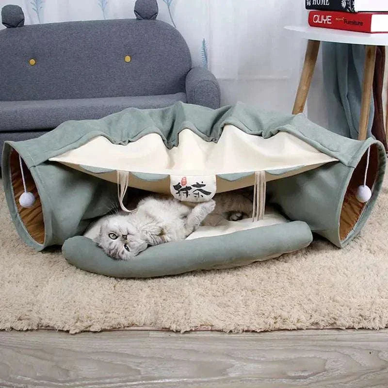 Cat tunnel bed in green and beige with hanging toys and a gray cat lying inside on a soft mat in a cozy living room setting.