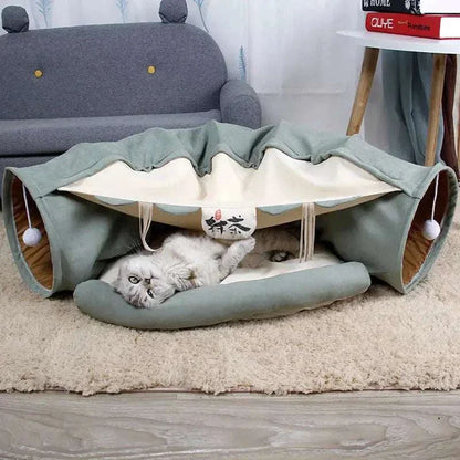 Cat tunnel bed in green and beige with hanging toys and a gray cat lying inside on a soft mat in a cozy living room setting.